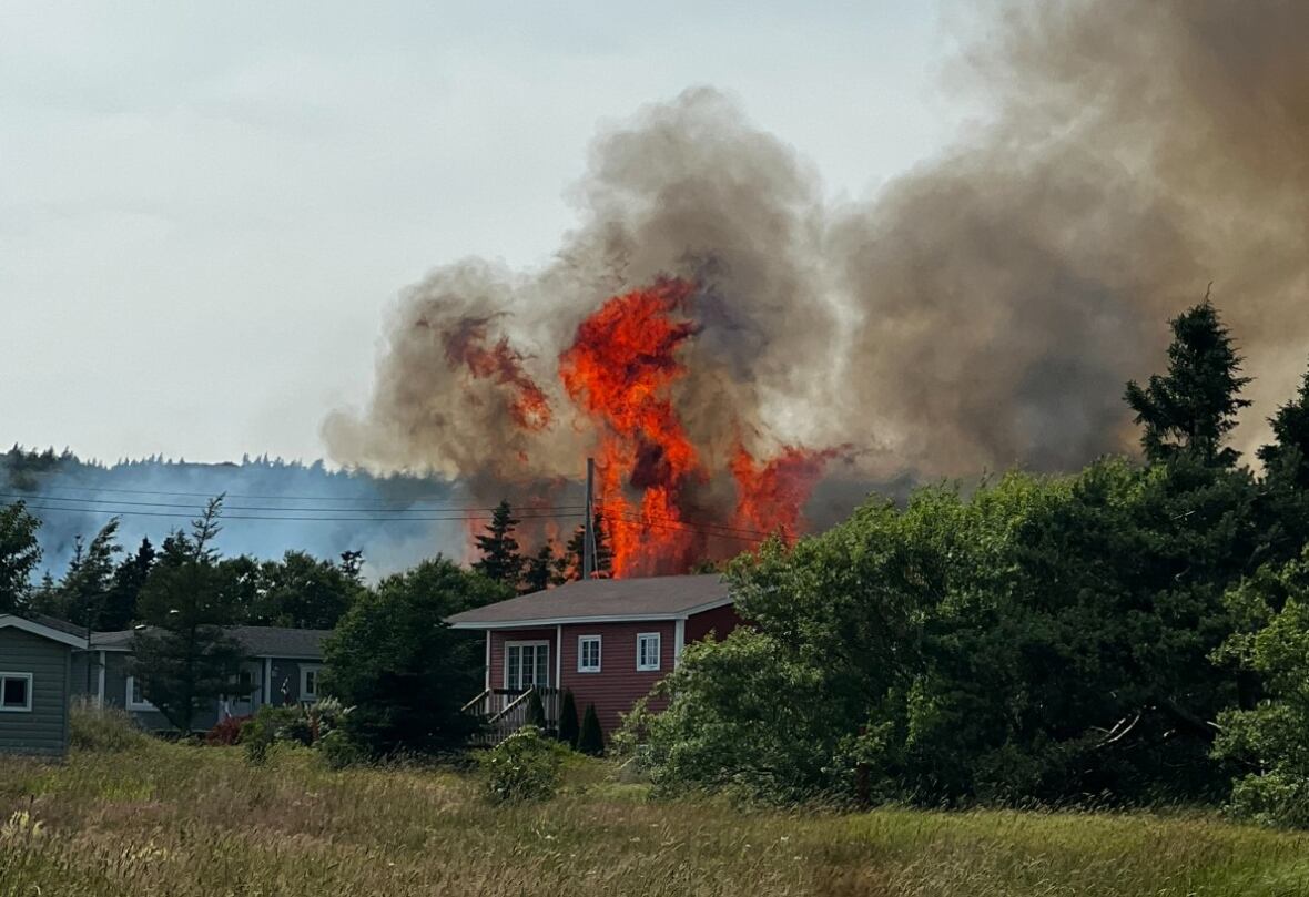 Large flames and plumes of black smoke billow from behind a small house in a grassy area.