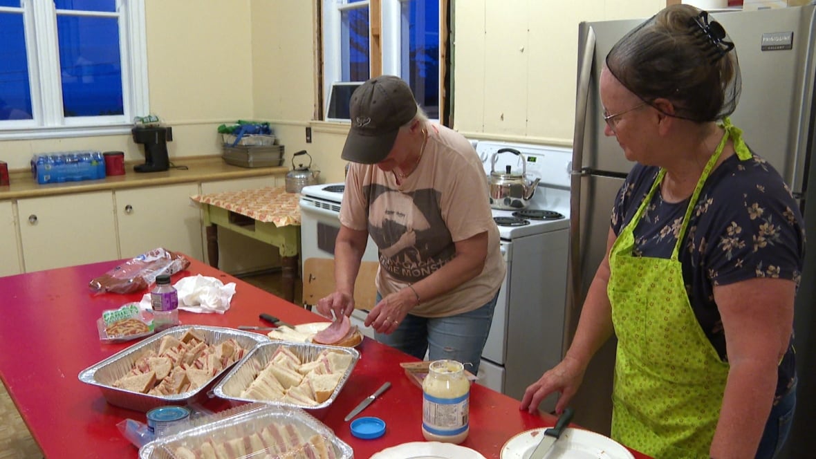 Two women stand in a kitchen making cold cut sandwiches with mayonnaise and white bread.