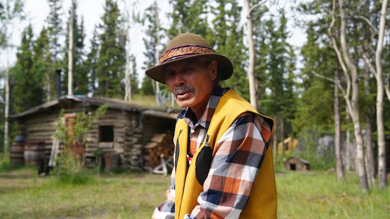 A man sitting, looking at the camera with a cabin in the background.