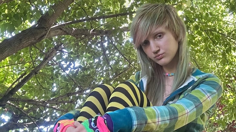 A person with long, asymmetrical, multi-coloured hair sits in front of a tree looking at the camera with their arms wrapped around their legs. 