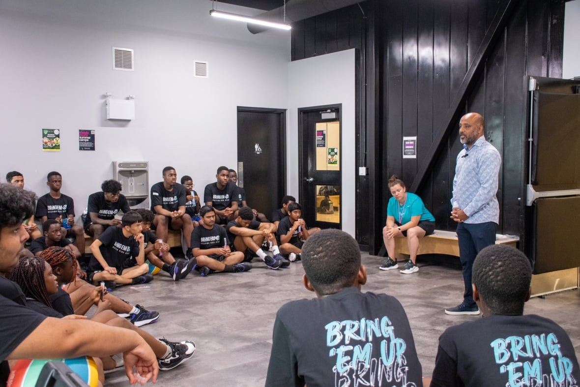 A group of diverse young people in matching black T-shirts gather in a circle around a man talking at the front. 