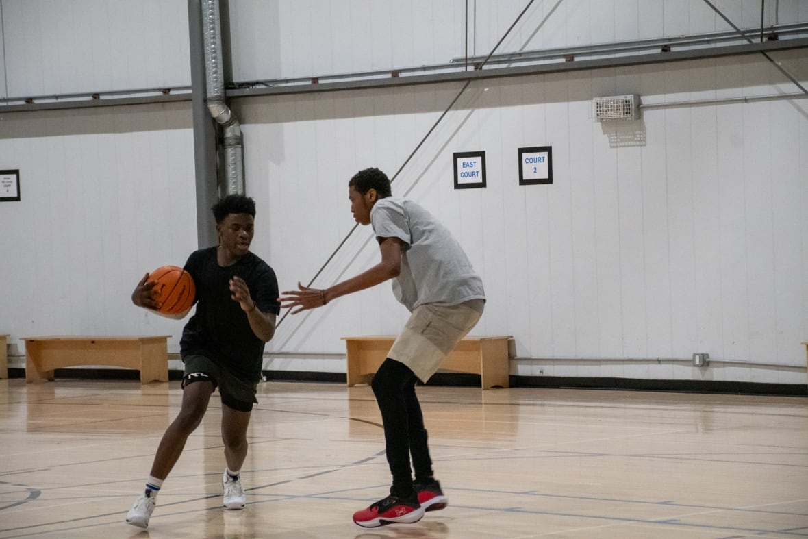 A young Black man in a black T-shirt tries to dribble around a taller defender.