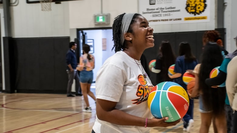 A smiling young girl in a white T-shirt holds a basketball in the middle of an indoor court.