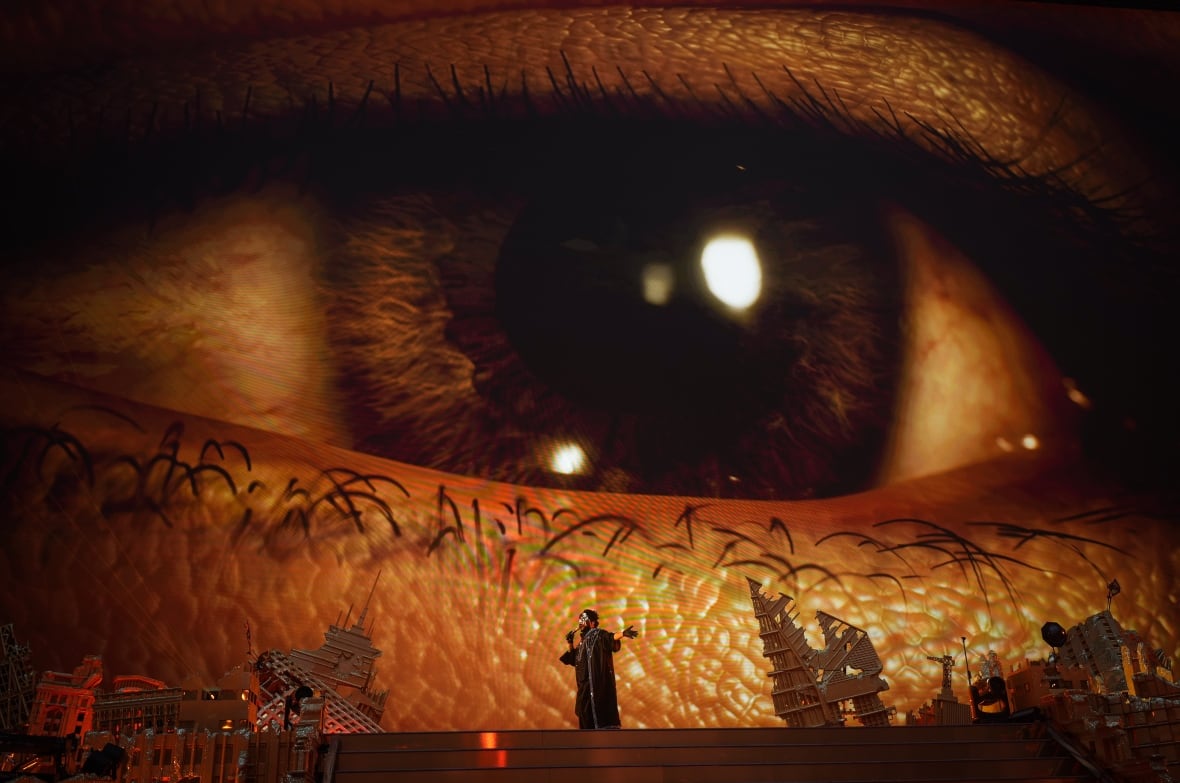 A picture of a human eye is projected behind a man singing into a microphone on stage.