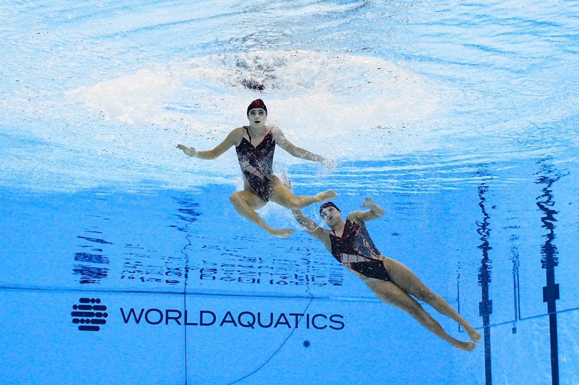 Two women wearing black swimsuits swim underwater in a pool.