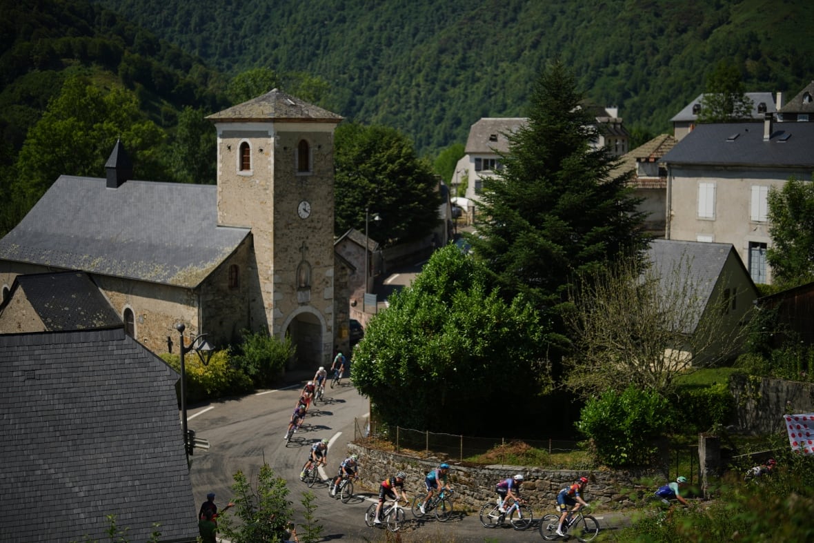 A group of people riding bikes ride through a street near stone buildings and green trees.