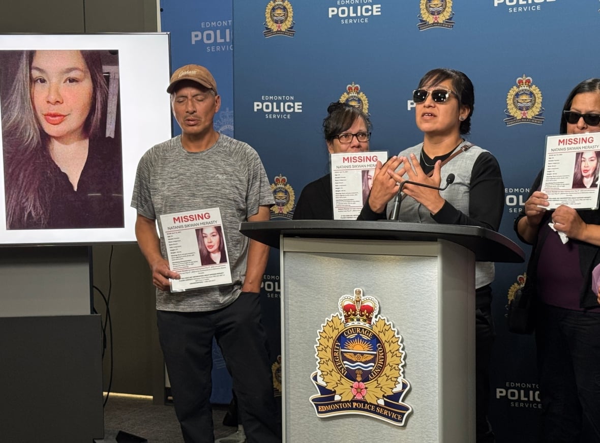 A woman wearing sunglasses stands at a podium surrounded by other people holding "missing signs" and a woman's face on a screen behind them.