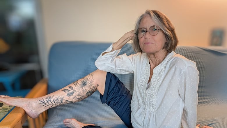 A woman with short grey hair smiles as she sits on a couch. Her right leg is propped up to show off a floral tattoo.