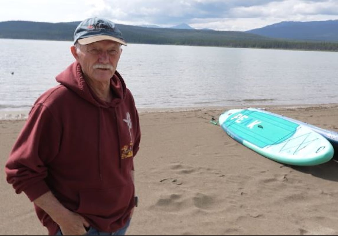 An older man stands on a sandy beach near a stand-up paddleboard.