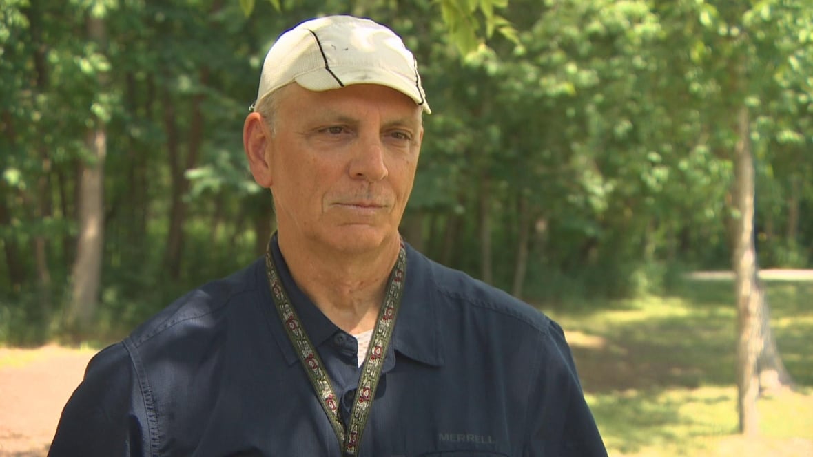 A man in a navy blue shirt and white hat speaks in front of a forested area.