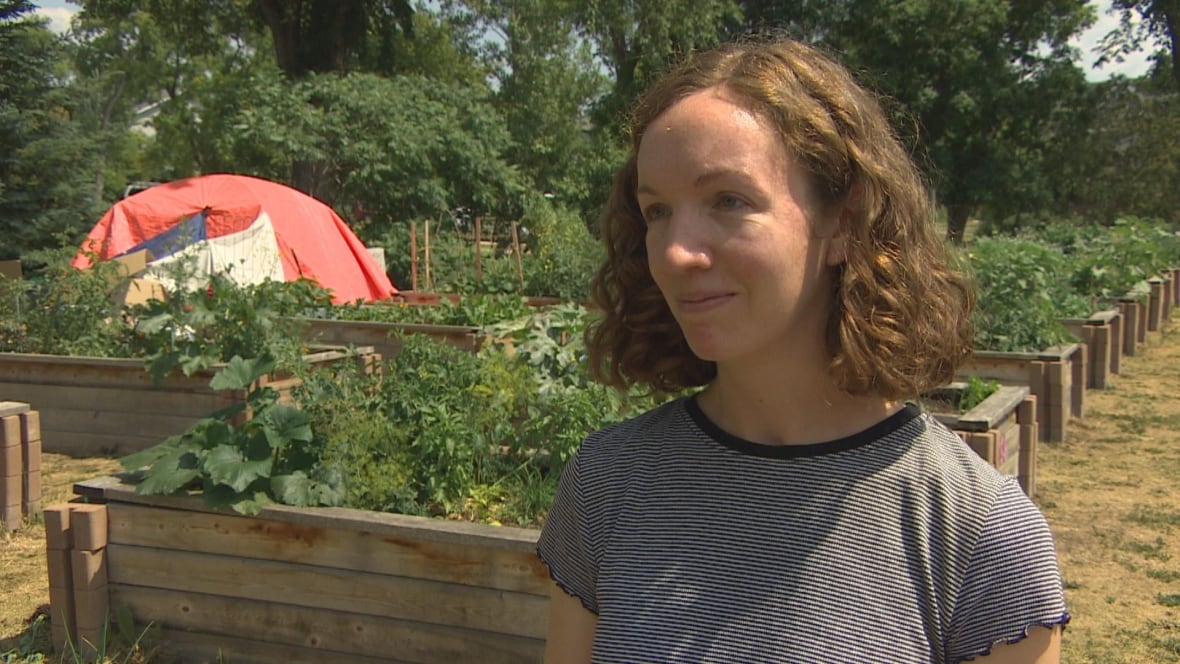 A woman with shoulder-length brown hair in a t-shirt stands in front of a community garden.