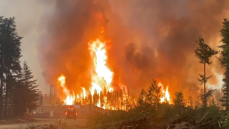A massive flame flares up in a wooded area behind a building and vehicle.