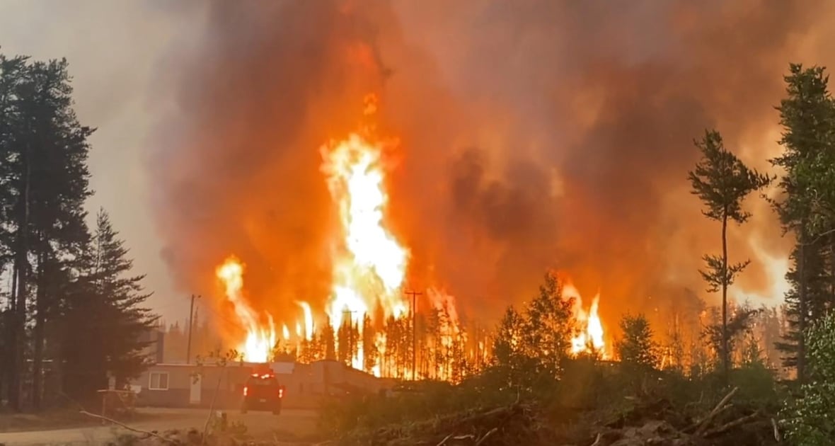 A massive flame flares up in a wooded area behind a building and vehicle.