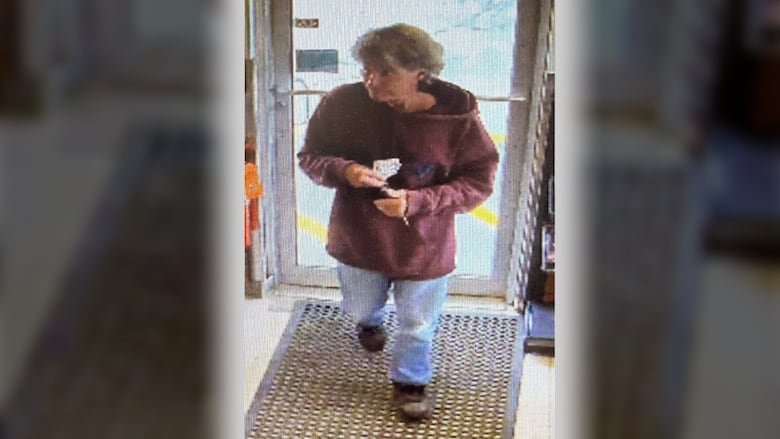 A man in a maroon hoodie and blue jeans walks into a store as seen through surveillance camera footage.