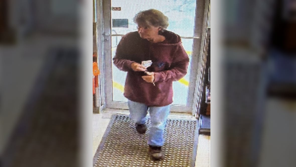 A man in a maroon hoodie and blue jeans walks into a store as seen through surveillance camera footage.