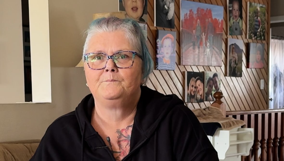 A white woman, with large glasses and short grey hair, is wearing a black button-up shirt. She is sitting down, straight-faced, inside a home. Collections of photos and art hang on the walls behind her.