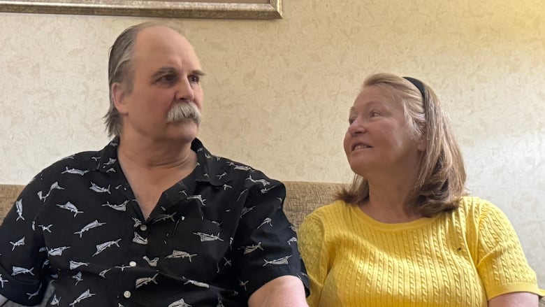 A balding white man, with a thick grey mustache, is wearing a navy blue button-up t-shirt. He is sitting on a couch, beside a white woman with red hair, who is wearing a bright yellow t-shirt. She is looking lovingly at him.