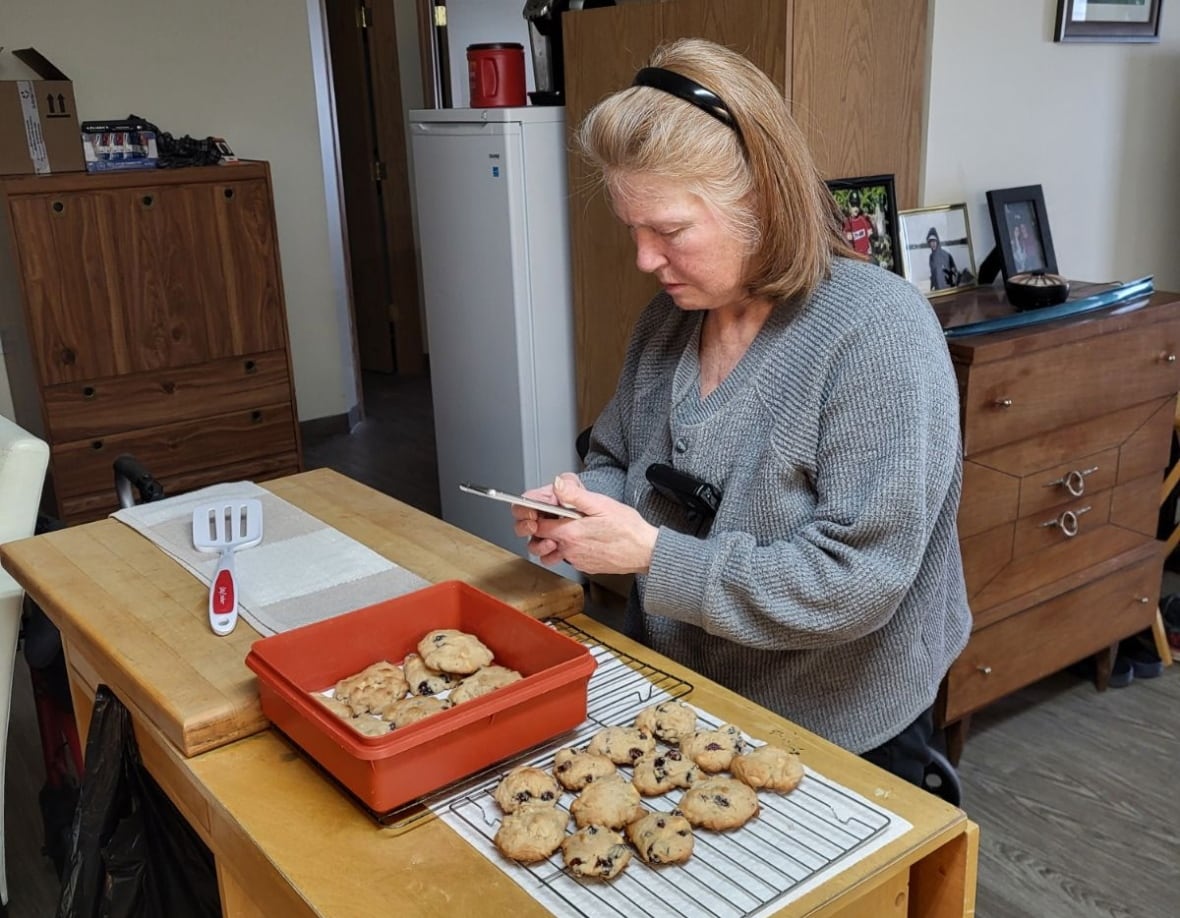 A white woman with red hair is wearing a grey sweater, while packing homemade cookies into a red container.