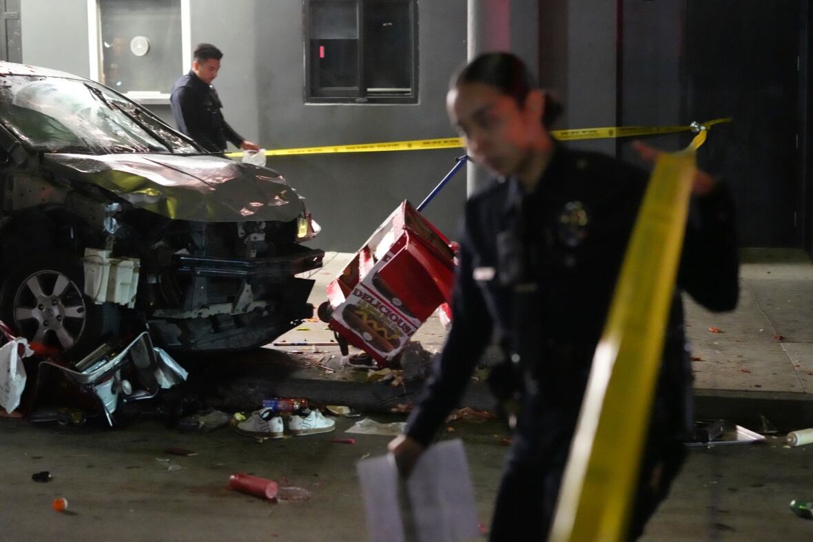 Law enforcement officers work around a small, damaged grey car covered in dents and debris.