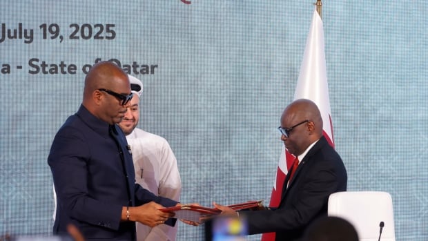 Three men stand on a stage, with two exchanging documents while the third looks on with a smile.