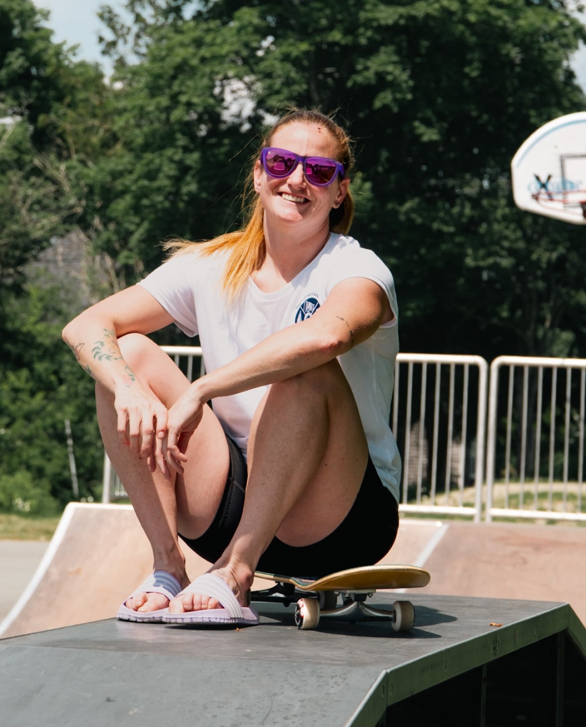 A white woman with blonde hair, wearing shades, sitting in front of a basketball net.