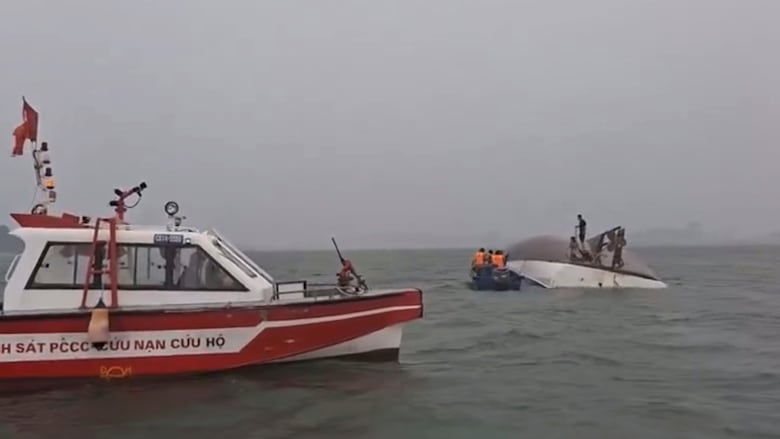 A boat is seen capsized with people standing on the overturned hull, while a larger boat idles nearby.