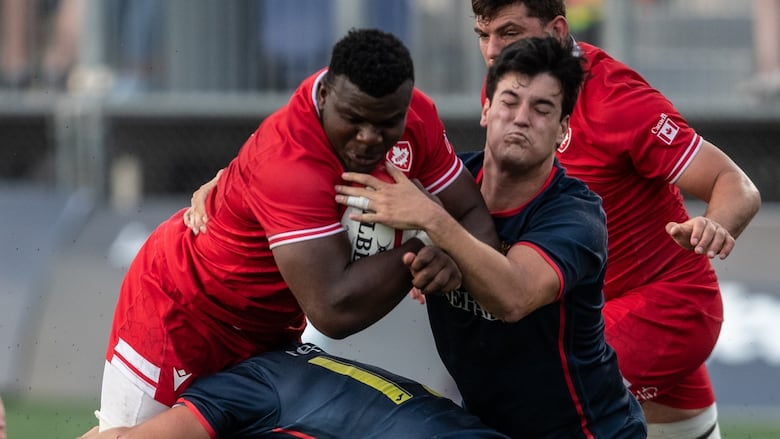 A male rugby player representing Canada is tackled by two Spanish players while carrying the ball in his right arm.