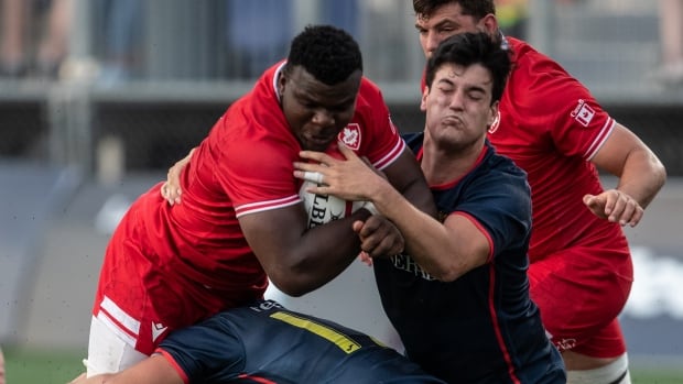 A male rugby player representing Canada is tackled by two Spanish players while carrying the ball in his right arm.