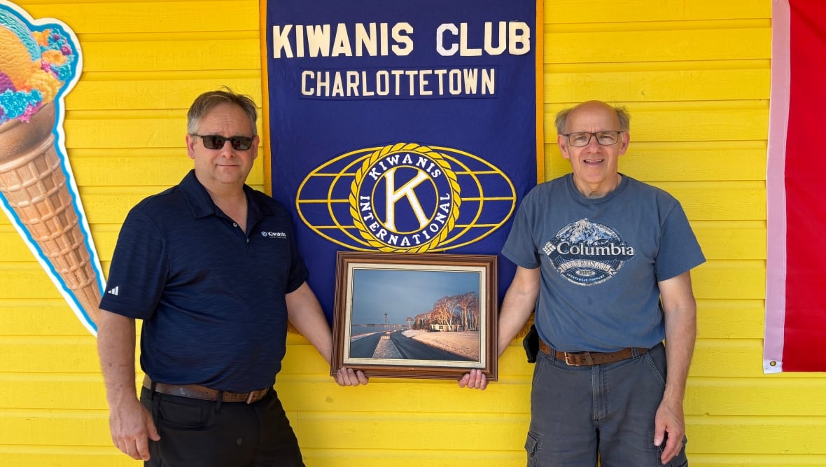 Two men with blue t-shirts hold a picture of the Kiwanis Dairy Bar, taken sometime between 1980 and 1990. Above the picture is a banner that reads "KIWANIS CLUB CHARLOTTETOWN"