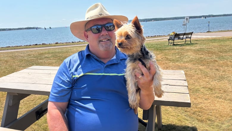 A man in a blue golf shirt, and sunhat sits at a picnic table, holding a dog in his left hand.