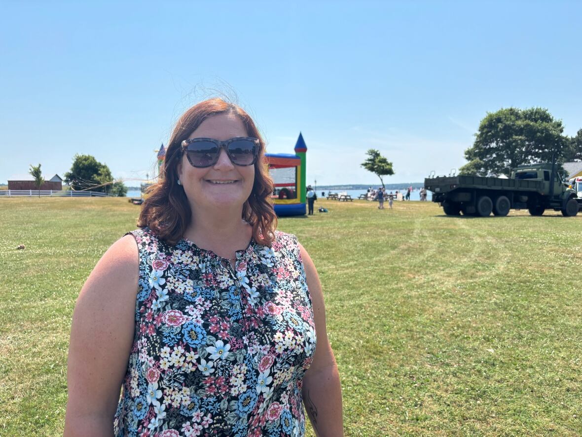 A woman in a flower print dress, wearing sunglasses stands in a field. Behind her is a bouncy castle and an army truck.