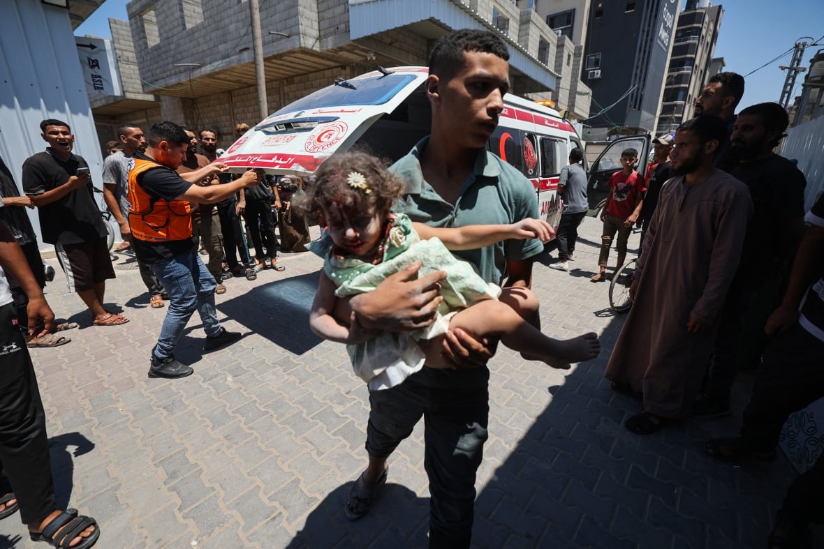 An adult carries an injured child covered in dust and blood into a hospital.