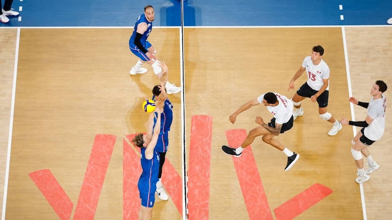A male volleyball player leaps into the air while hitting the ball over two opponents who are attempting to block it at the net.