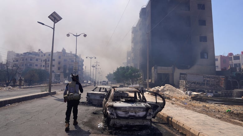 An armed fighter stands near destroyed vehicles as a smoldering building is seen behind.