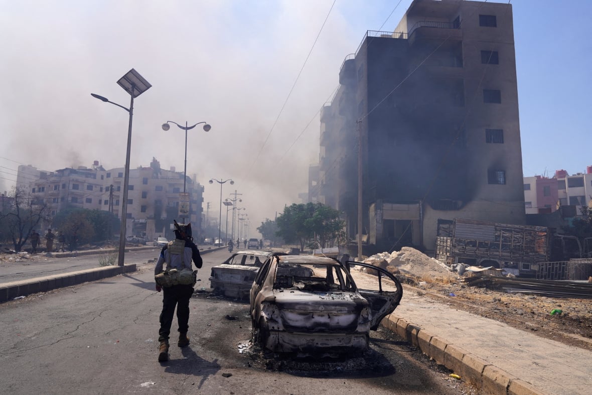 An armed fighter stands near the destroyed vehicles where a burning building appears.