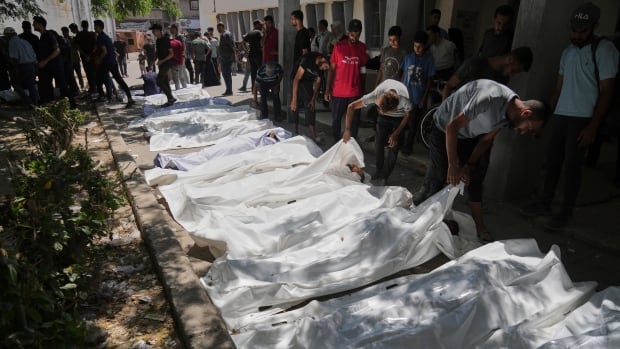 Several bodies covered in white plastic are laid on the street as people peek beneath to see if it is their loved ones.