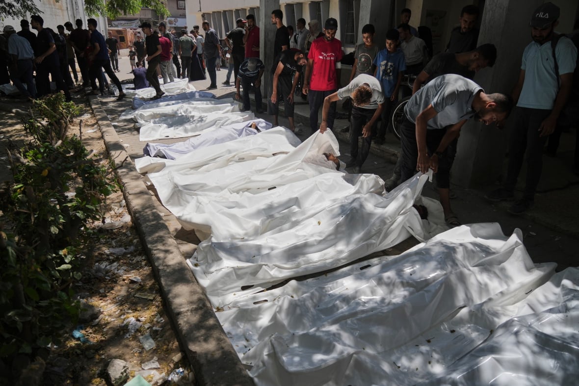Several bodies covered in white plastic are laid on the street as people peek beneath to see if it is their loved ones.