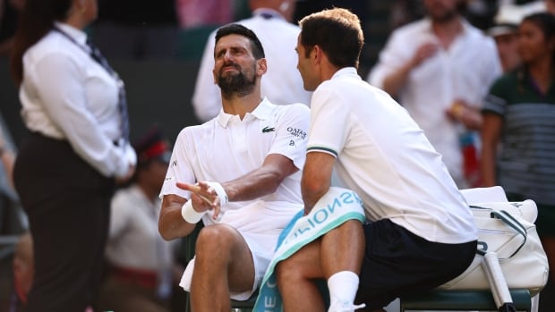 A male tennis player winces in pain while receiving medical attention on his left groin during a break in a match.