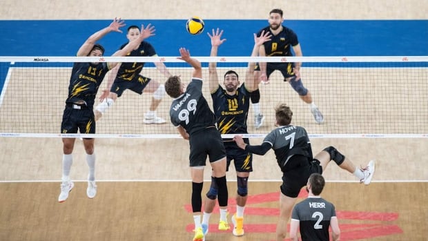 A male volleyball player representing Canada hits the ball over the net as an opponent representing Ukraine attempts to block with his arms in the air.