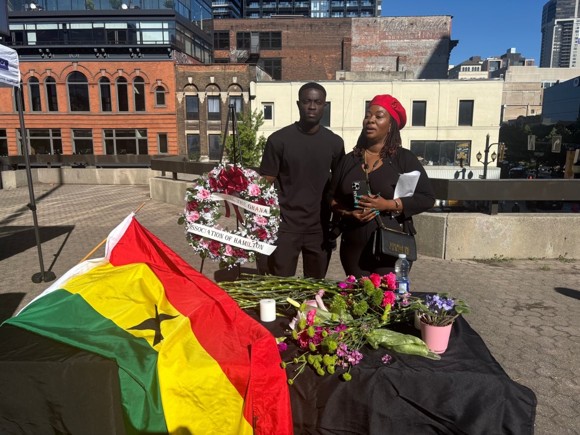 A woman speaks while a man stands next to her. A Ghanaian flag is set on a table in front of them next to several flowers.