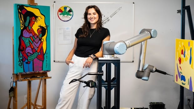 A woman leans on a robotic arm next to two paintings.