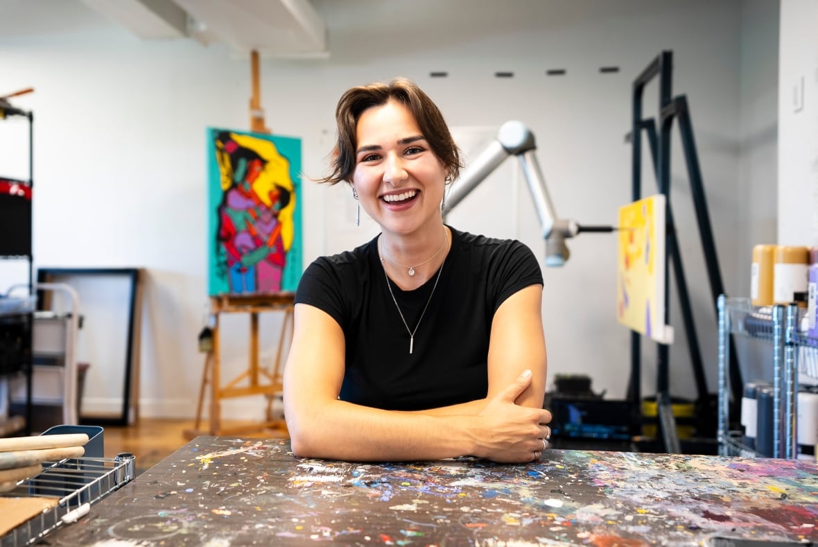 A woman smiles while on a painter's table, with paintings visible behind her.
