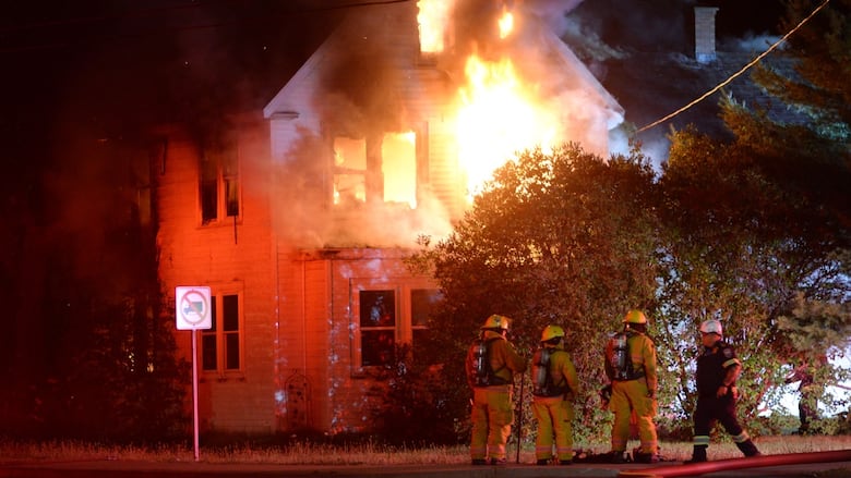 Four firefighters can be seen standing in front of a two and a half story house that is on fire. 