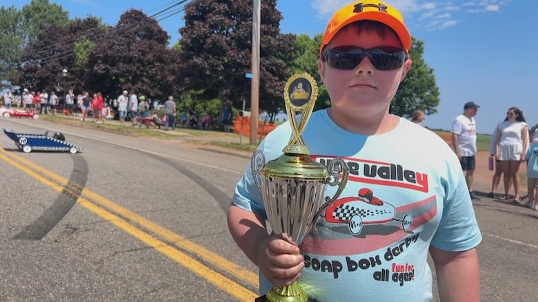 A boy in a orange baseball cap and blue t-shirt looks at the camera holding a gold trophy.