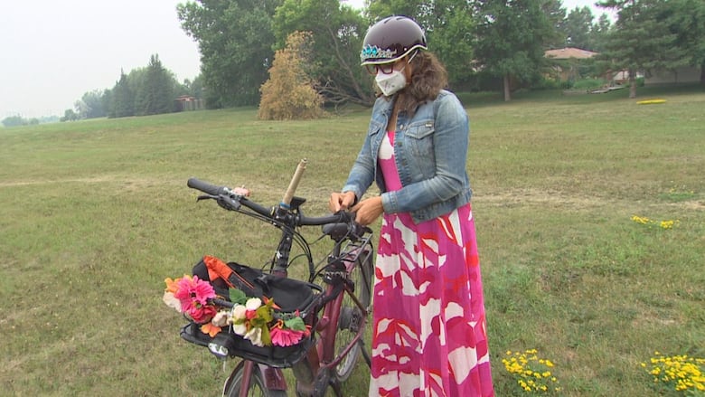 A woman wearing a face mask, red dress and bicycle helmet is seen with her bicycle.