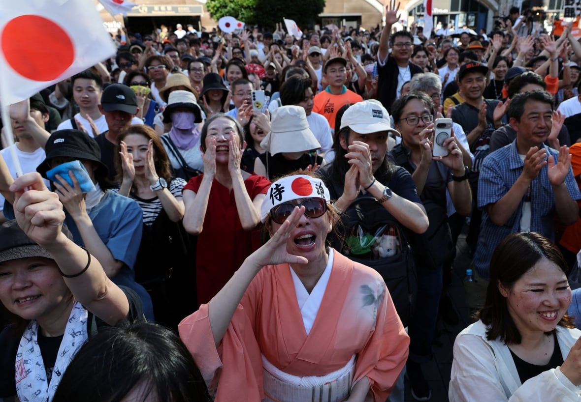 An outdoor crowd of dozens of Asian people is shown. One wears a headband and martial arts type robe, while a Japan flag is also shown.