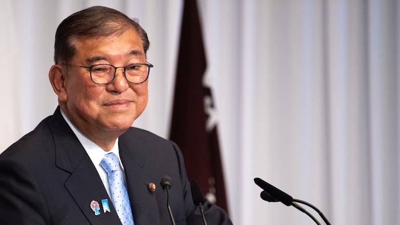 A cleanshaven, bespectacled older Asian man wearing suit and tie speaks into a microphone at a podium.