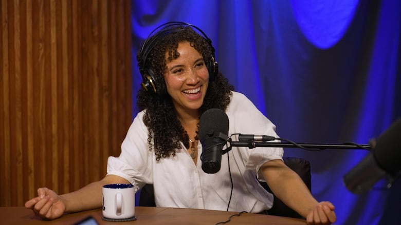 A smiling woman wearing over-ear headphones while sitting in front of a studio microphone.