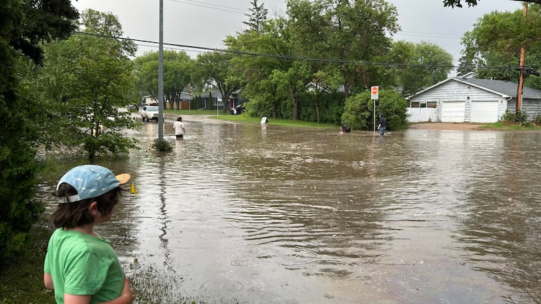 A couple of people wade into flooded residential streets on a cloudy day.