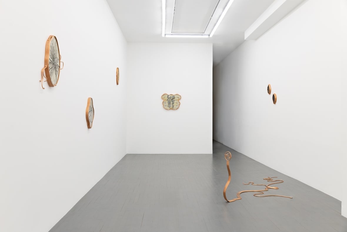 A white-walled exhibition space is hung with stained glass artworks framed by copper. On the floor is a large, snakelike copper sculpture.